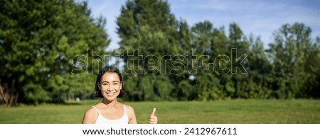 Vertical shot of asian woman shows thumbs up, recommending yoga training online, meditation app, doing exercises on fresh air in park.