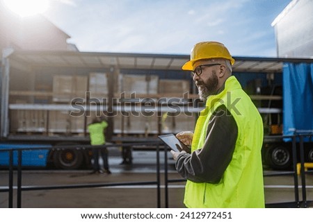 Warehouse manager overseeing unloading of truck, holding tablet, looking at cargo details, checking delivered items, goods against order. Royalty-Free Stock Photo #2412972451