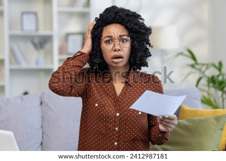 Worried young woman with glasses holding a document, expressing stress and confusion at home, concept of financial or personal crisis. Royalty-Free Stock Photo #2412987181