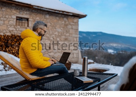 Mature man working from cozy cabin in mountains, sitting on terrace with laptop, enjoying cup of coffee. Concept of remote work from beautiful, peaceful location. Hygge at work.