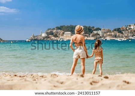 Woman and her little daughter in swimsuit standing together on the beach, watching at the Port de Soller bay in Mallorca. Holidays with children concept. Royalty-Free Stock Photo #2413011157