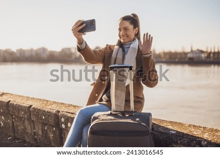 Happy woman  tourist with suitcase taking selfie  and waving while resting by the river. Toned image.