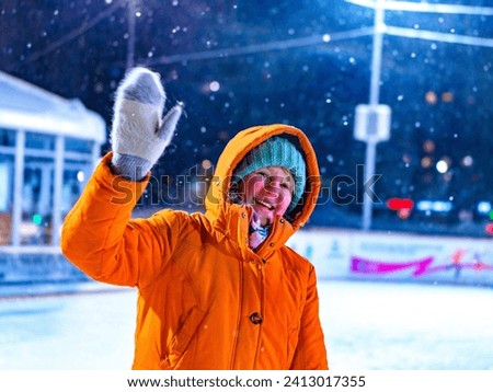 A young woman walking on an ice rink smiles cheerfully and raises her hand in greeting. Setting of winter evening and snowfall.