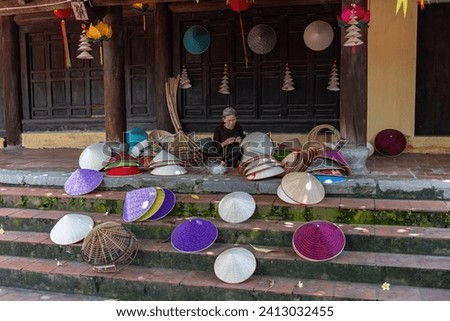 Focus an old woman making conical hats by hand in front of temple - Vietnamese name is Non la. Lang Chuong village, Hanoi city, Vietnam. Travel concept. Royalty-Free Stock Photo #2413032455