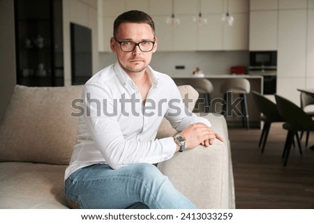 Handsome man relaxing on couch in his apartment. Young male looking at camera with serious expression on face. Portrait of man in casual clothes and glasses sitting on background of modern kitchen.