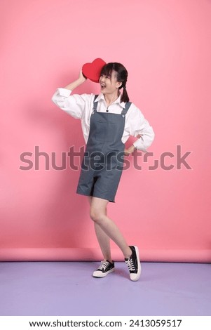 Cheerful lovely young asian woman in overalls or casual clothes with gesture of holding red heart-shaped isolated on pink background. St Valentine's Day, Women's Day, Birthday
