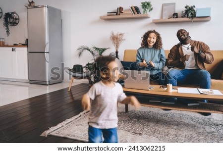 Cheerful young couple in casual clothes sitting on sofa and cheering cute little girl running and playing around in living room while spending time together