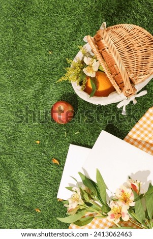 A bamboo basket container fruits and flowers decorated with white notebooks and checkered fabric on a green grass background. Top view, copy space for advertising. Summer concept