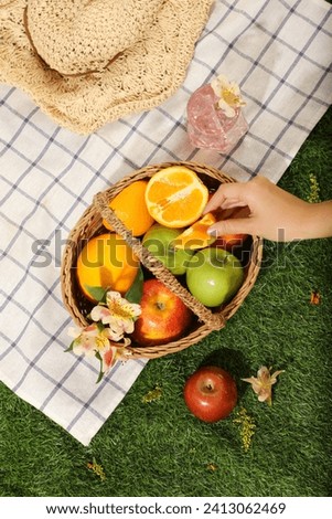 Top view of bamboo basket full with fruits: oranges, apple and green apples. A hand is holding an orange slices. Hat, cool of water cup and checkered decorated for picnic day