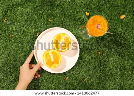 On a green grass background, a hand of woman is placing a white plate containing delicious orange slices next to a cool smoothie. Top view, minimal scene for advertising with summer concept