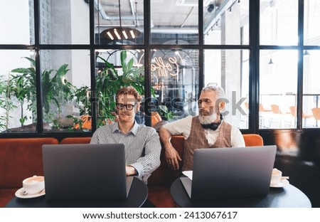Concentrated male freelancers browsing laptop while sitting at table with cup of coffee and working remotely in modern workspace near big potted plants on background
