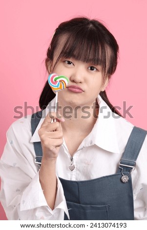 Cheerful lovely young asian woman in overalls casual clothes with gesture of holding heart shaped lollipop isolated on pink background. St Valentine's Day, Women's Day, Birthday, Sweet tooth concept