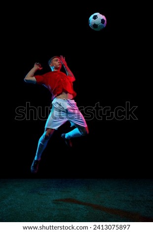 Airborne Mastery. Portrait of soccer as professional player executes flawless mid-air kick, sending ball soaring against black background in neon light. Concept of sport games, hobby, energy. movement