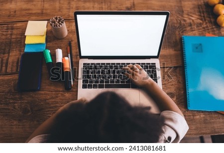 Overhead view of a child's hands typing on a laptop computer with a blank screen, surrounded by colorful study materials on a wooden desk, depicting a home learning scene Royalty-Free Stock Photo #2413081681