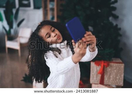 Portrait of cute African American female kid with long curly hair and casual clothes making round mouth while taking selfie on mobile phone and standing in living room against blurred background