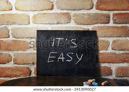 Black board with the phrase IT'S EASY drown by hand on wooden table on brick wall background. Presentation concept.