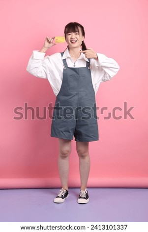 Cheerful lovely young asian woman in overalls casual clothes with gesture of holding golden bank credit card on pink background. Valentine's Day, Women's Day, Birthday, Casual Outfits