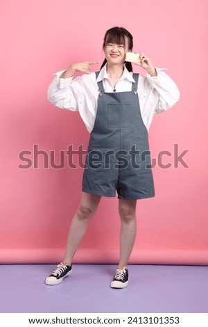 Cheerful lovely young asian woman in overalls casual clothes with gesture of holding golden bank credit card on pink background. Valentine's Day, Women's Day, Birthday, Casual Outfits