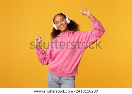 Happy African American teenager student girl enjoying music with earphones, dancing with eyes closed and raised arms, on yellow background. Musical playlist and favorite songs concept