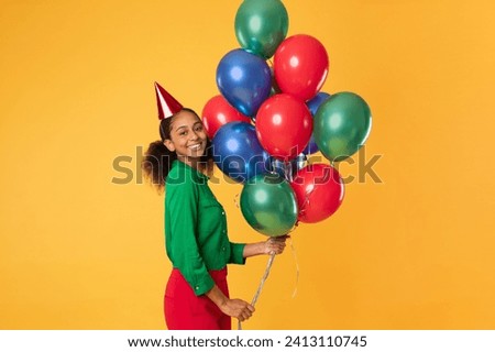 Happy black adolescent teen girl in birthday hat holding bunch of colorful balloons, smiling to camera, posing over yellow studio backdrop. Bday celebration and party concept