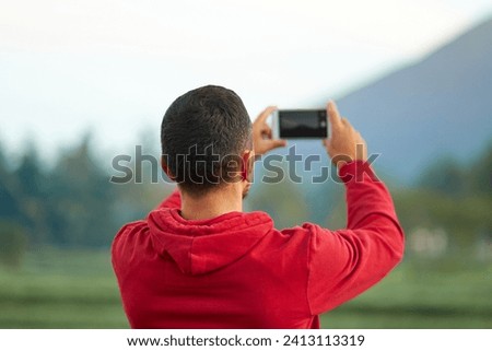 A young man stands on the side of the road near a rice paddy and takes a picture with his phone of Mount Agung on the popular tourist island of Bali
