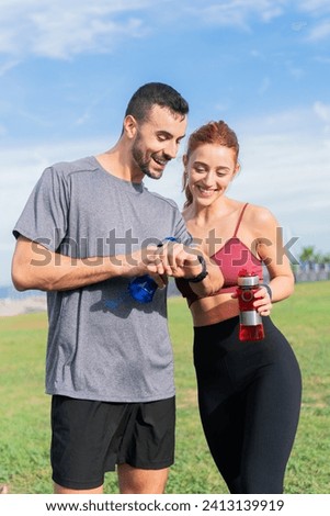 Two sporty young adults resting and checking the smart band analyzing the workout smiling and laughing together outdoors. Two people fitness resting with water bottles and with sportswear Royalty-Free Stock Photo #2413139919