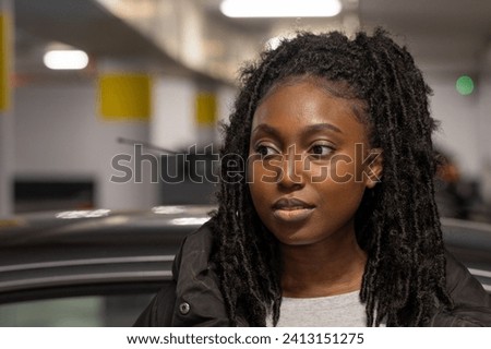 In this image, a young woman is pictured in a candid, side-profile view against the backdrop of an indoor parking lot. The focus is on her as she appears lost in thought, with her textured black