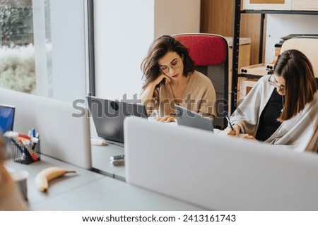 Female businesspeople collaborating and discussing project solutions in a modern office lobby. They are happy and focused as they work together on tasks using technology. Royalty-Free Stock Photo #2413161743