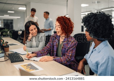 Diverse colleagues working in a call center. Focus on a hispanic woman call center agent answering incoming calls with a headset and writing notes while providing customer service remotely. Royalty-Free Stock Photo #2413164881
