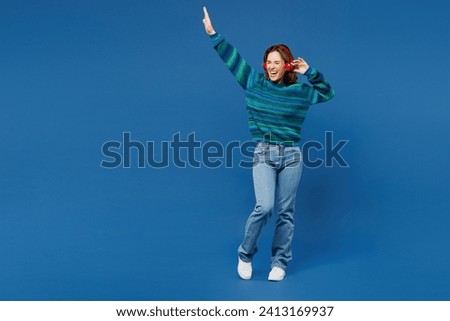 Full body happy young woman she wears knitted sweater casual clothes listen to music in headphones raise up hands dance isolated on plain blue cyan color background studio portrait. Lifestyle concept
