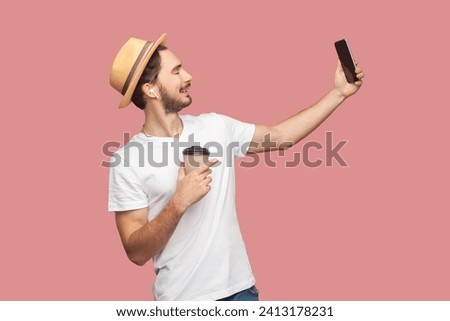 Side view portrait of smiling handsome attractive bearded man in white T-shirt and hat standing holding takeaway coffee and making selfie on smartphone. Indoor studio shot isolated on pink background.