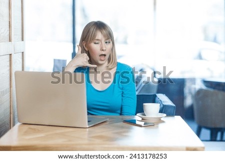 Portrait of flirting attractive young woman with blonde hair in blue shirt working on laptop, having video call, asking to call her back. Indoor shot in cafe with big window on background.
