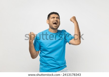 Portrait of unshaven man wearing blue T- shirt standing clenches fists with positive expression, rejoices wonderful news, smiles happily. Indoor studio shot isolated on gray background. Royalty-Free Stock Photo #2413178343