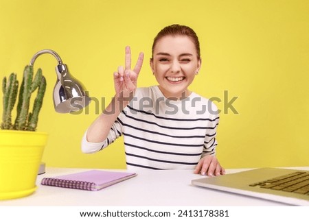 Beautiful excited woman looking at camera POV, point of view of photo, showing victory gesture, sitting at work place with laptop. Indoor studio studio shot isolated on yellow background.