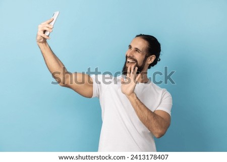 Portrait of man with beard wearing white T-shirt taking selfie and waving hand, gesturing hello for followers, communicating on video call. Indoor studio shot isolated on blue background.