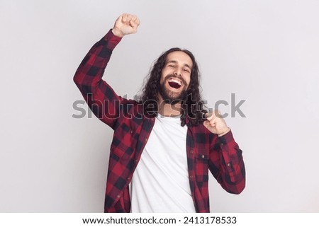 Cheerful triumphing man with long curly hair in checkered red shirt achieves victory, raises clenched fists with triumph, rejoices winning prize. Indoor studio shot isolated on gray background. Royalty-Free Stock Photo #2413178533
