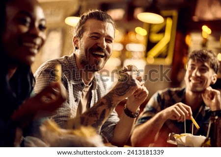 Cheerful man eating hamburger while gathering with friends in a bar. 
