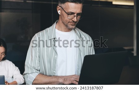 Caucasian businessman in optical eyewear and earbuds browsing website with marketing webinar, skilled male IT professional using bluetooth connection for listening audio via application on laptop