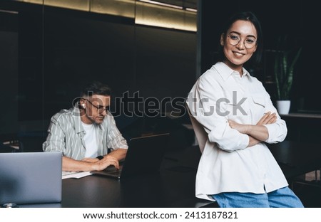 Half length portrait of happy Asian female employee with crossed hand smiling at camera while posing in coworking with male colleague on background, successful graphic designer in spectacles