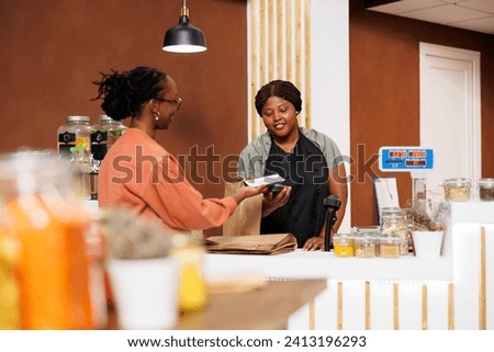 Young African American woman, shopping at a modern supermarket, browsing products, and paying with mobile device. The store owner operates the POS terminal at the checkout counter. Royalty-Free Stock Photo #2413196293