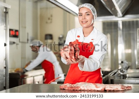 Smiling young girl in white uniform and red oilcloth apron working in meat processing room of butchery, standing near cutting table with fresh raw beef heart in hands.. Royalty-Free Stock Photo #2413199133