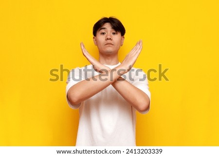 asian man in white t-shirt shows stop and prohibit gesture with crossed arms on yellow isolated background, korean guy rejects and warns about danger