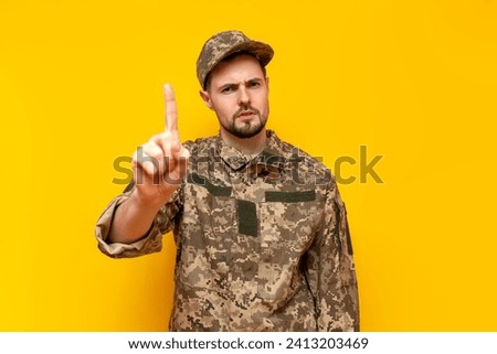 young Ukrainian male soldier in a camouflage pixel uniform warns and threatens on a yellow isolated background, guy military cadet of the Ukrainian army points with his finger
