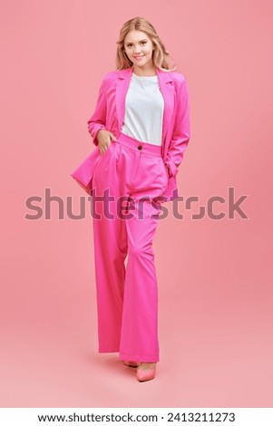 A beautiful girl with wavy blonde hair and gentle pink makeup, dressed in a pink pantsuit, poses on a pink studio background. Full-length portrait.
