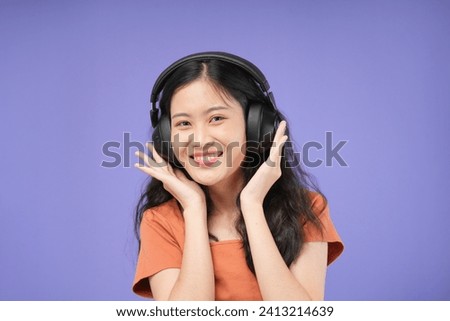 Happy positive Asian woman enjoys music uses wireless headphones holds turns away enjoys spare time gets pleasure from song wear orange shirt indoor copy space righ