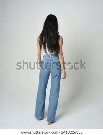 Full length portrait of brunette female model wearing casual clothes, white singlet shirt, denim jean pants. Standing pose walking away from camera. Isolated on white studio background. Royalty-Free Stock Photo #2413226355