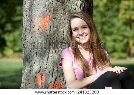 An attractive and happy young brunette girl sitting next to a tree on a sunny day.