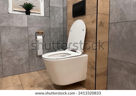 A modern bathroom with a ceramic toilet in white. The toilet is closed and has a seat and a flush. The bathroom has a wall and a floor with tiles. Royalty-Free Stock Photo #2413320833