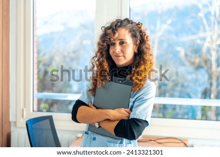 Tired nurse smiling holding medical reports and sitting on the desk next to a window in the hospital