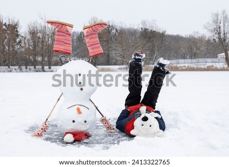 funny snowman in red boots stands upside down on frozen snowy lake. boy in polar bear hat lies next to him cheerfully raising legs. seasonal active recreation, carefree childhood. Holidays winter fun
